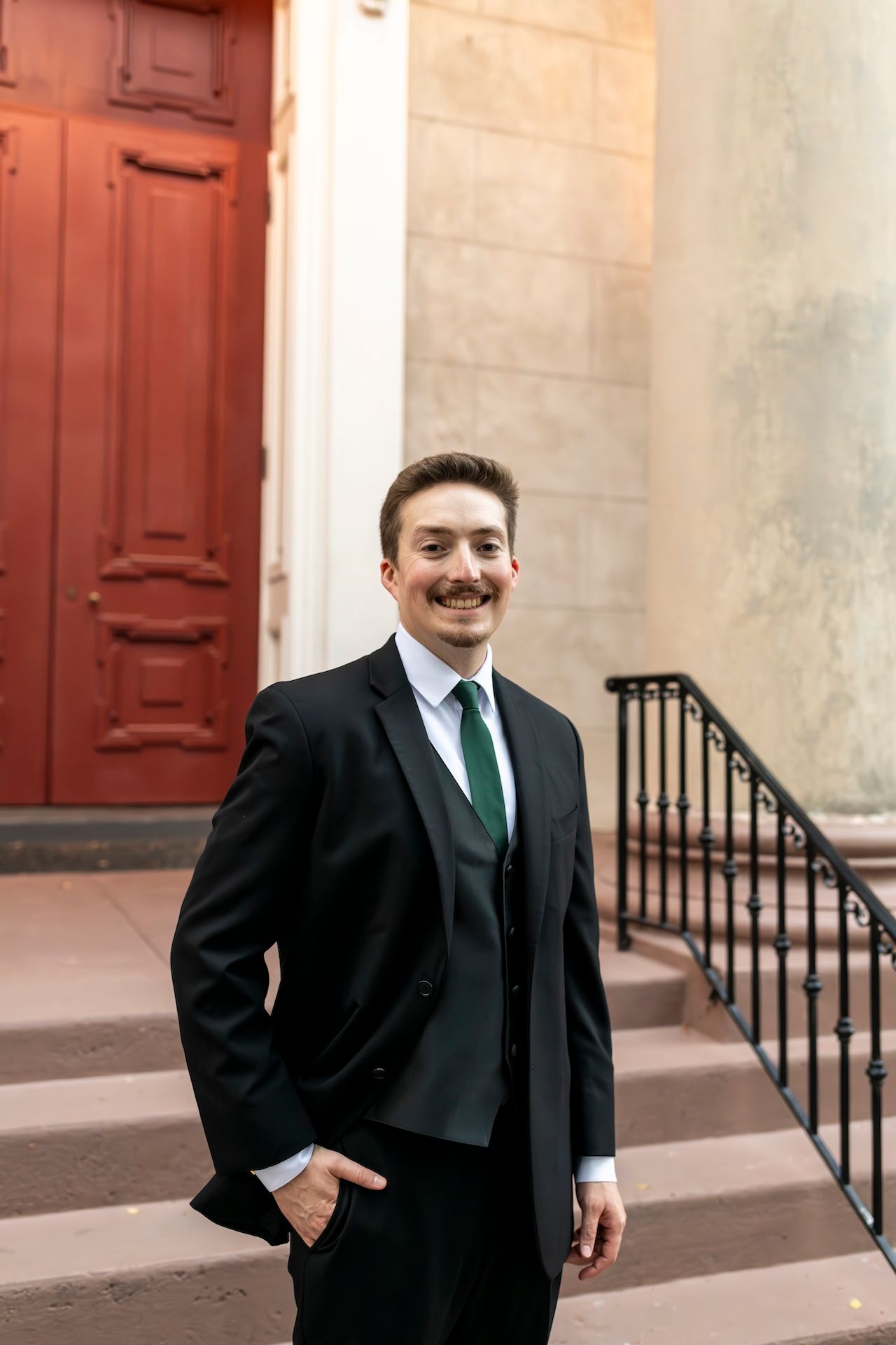 Smiling professional in a black suit and green tie stands by a brick building with a red door and iron railing.