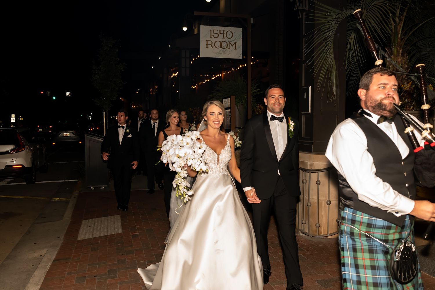 A celebratory wedding procession walks through a downtown area at night with a bagpiper in traditional Scottish attire.