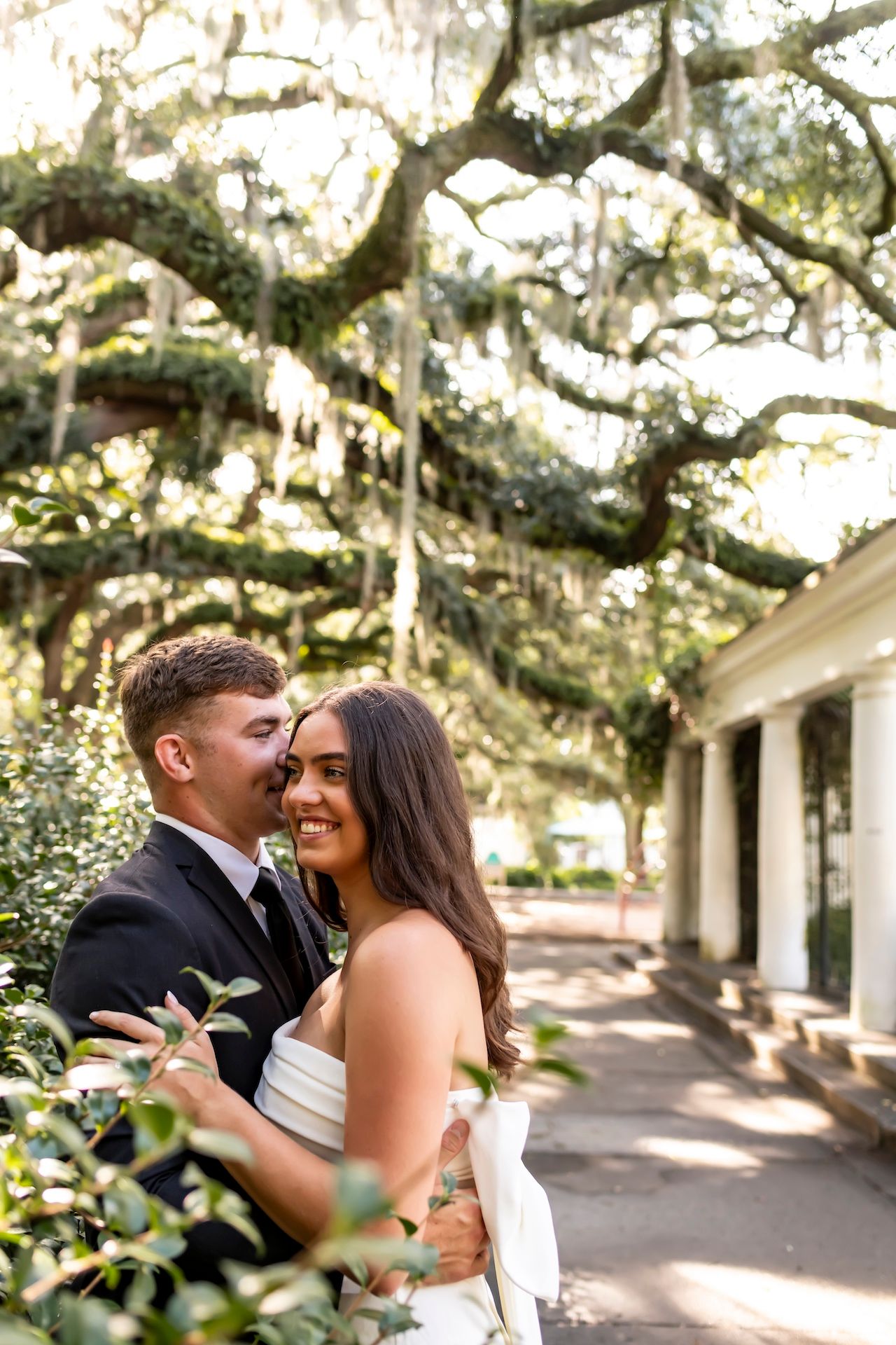 Couple embraces intimately under a canopy of moss-draped oak trees and white pillars in dreamy outdoor setting.
