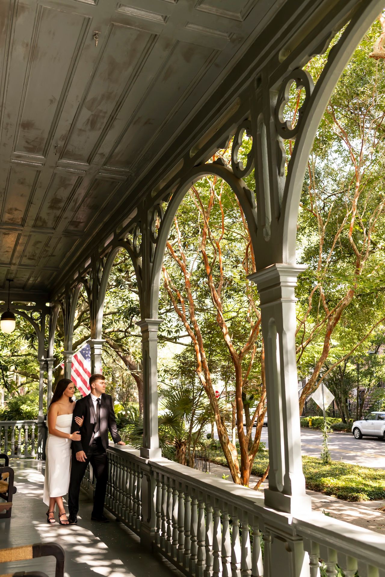 Two people stand on a historic southern-style porch with ornate white columns and archways framed by green trees.