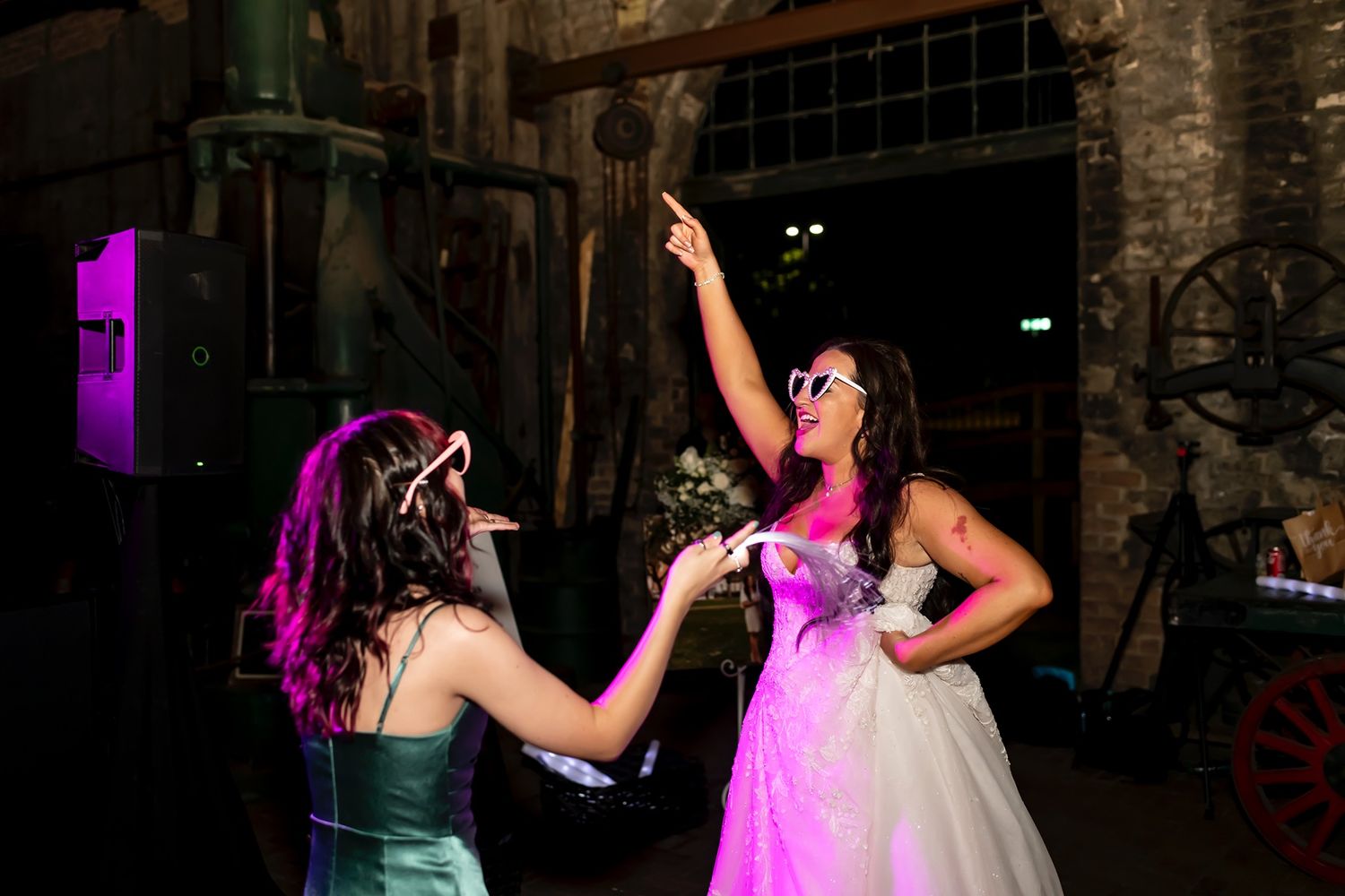 Two people in formal dresses dance together under atmospheric lighting at an event venue with stone walls.