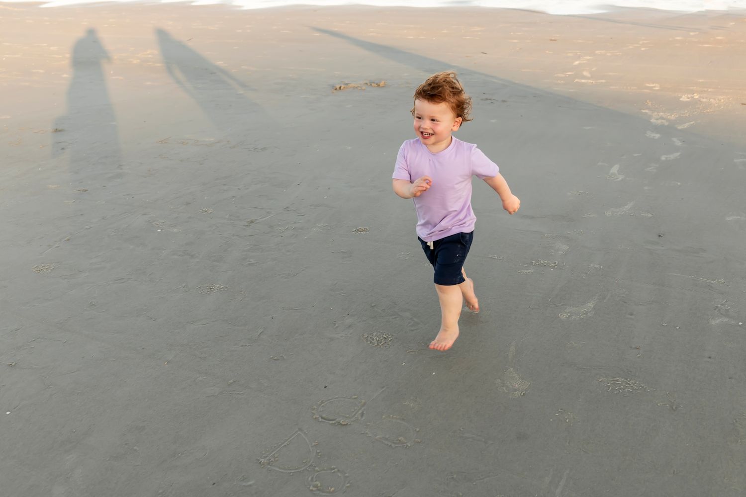 A joyful young person runs barefoot on a sandy beach at sunset, wearing a pink shirt and dark shorts.