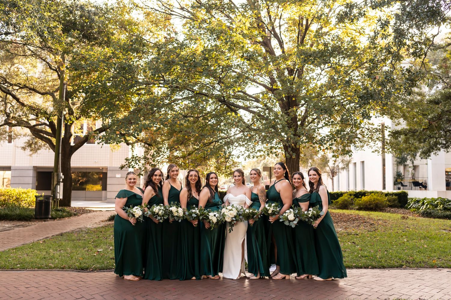 Wedding party wearing emerald green bridesmaid dresses with white bouquets poses outdoors on a brick walkway under trees.