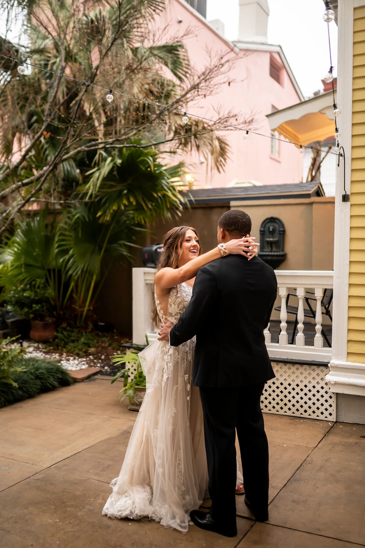 Couple embraces romantically in front of a pink historic building with tropical landscaping on a patio.