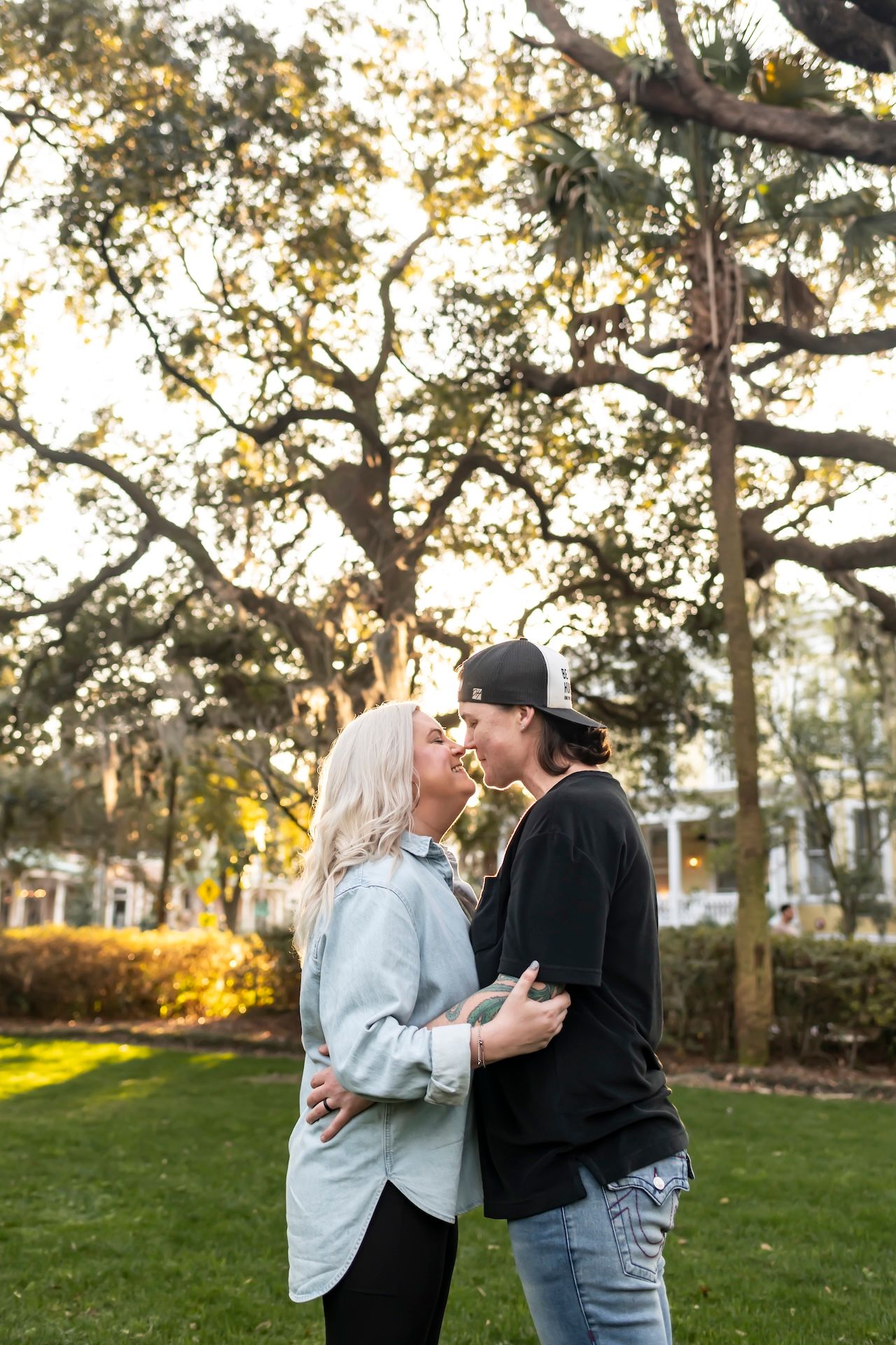 A couple shares a romantic moment outdoors in a park during sunset with tall trees in the background.