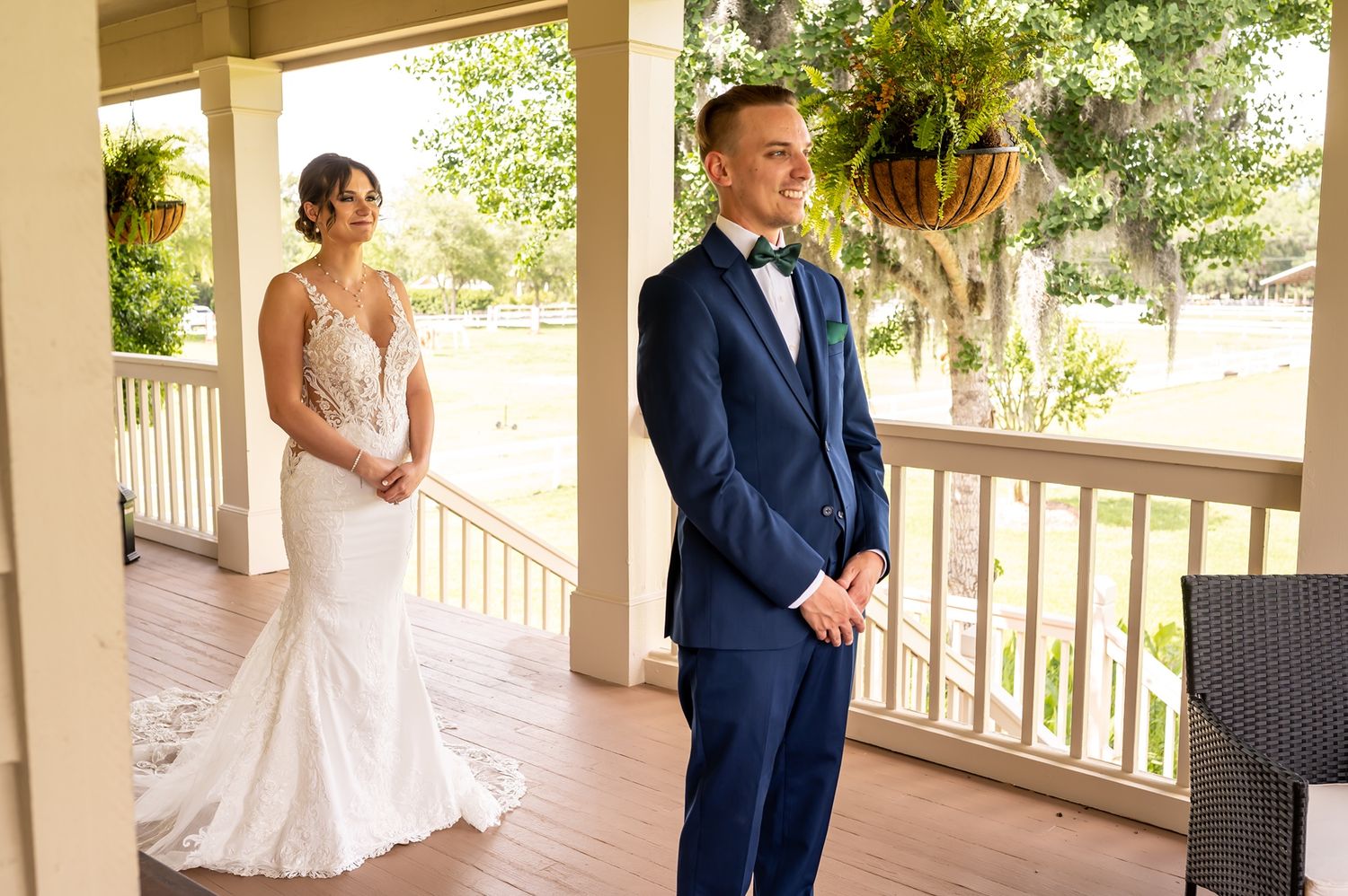 A couple shares a tender first look moment on a covered porch with hanging planters in the background.
