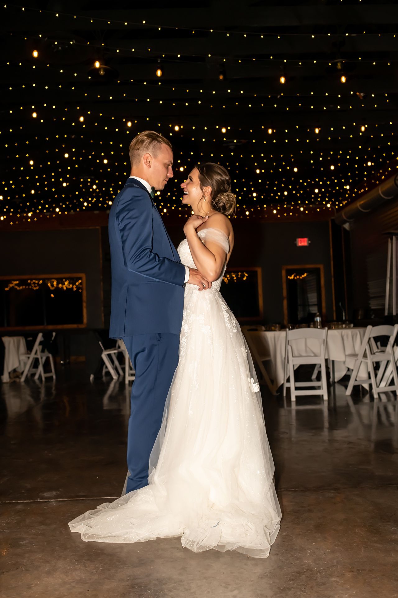 A couple in wedding attire share a romantic moment on their first dance under twinkling string lights in an elegant venue.