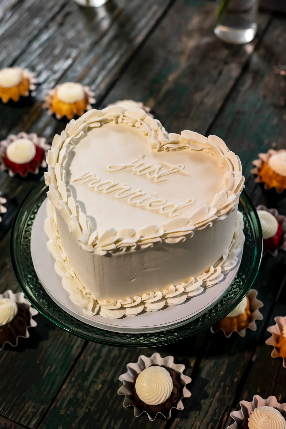 Heart-shaped white frosted cake with swirled borders displayed on black plate against rustic wooden surface with cupcake wrappers.
