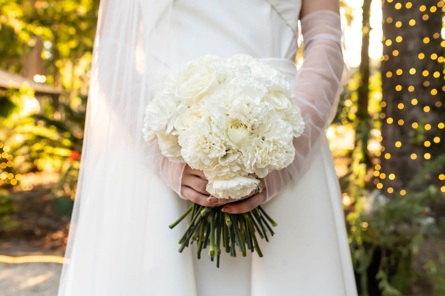 A bridal bouquet of white peonies and roses held by gloved hands with twinkling lights in background.