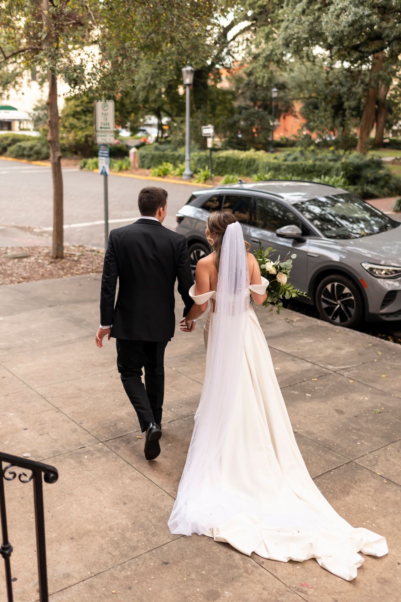 Newlyweds walking together on a sidewalk with the bride wearing a flowing white dress with long veil and groom in black suit.