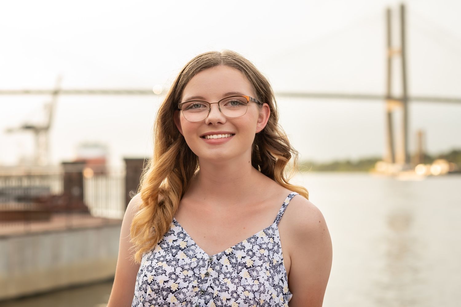 A person in a floral sundress stands smiling by the waterfront with a bridge visible in the background.