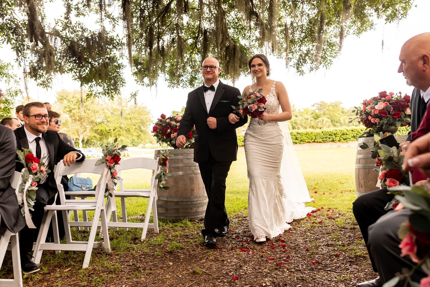 Two people walk down the grassy aisle at an outdoor wedding ceremony under Spanish moss draped trees.