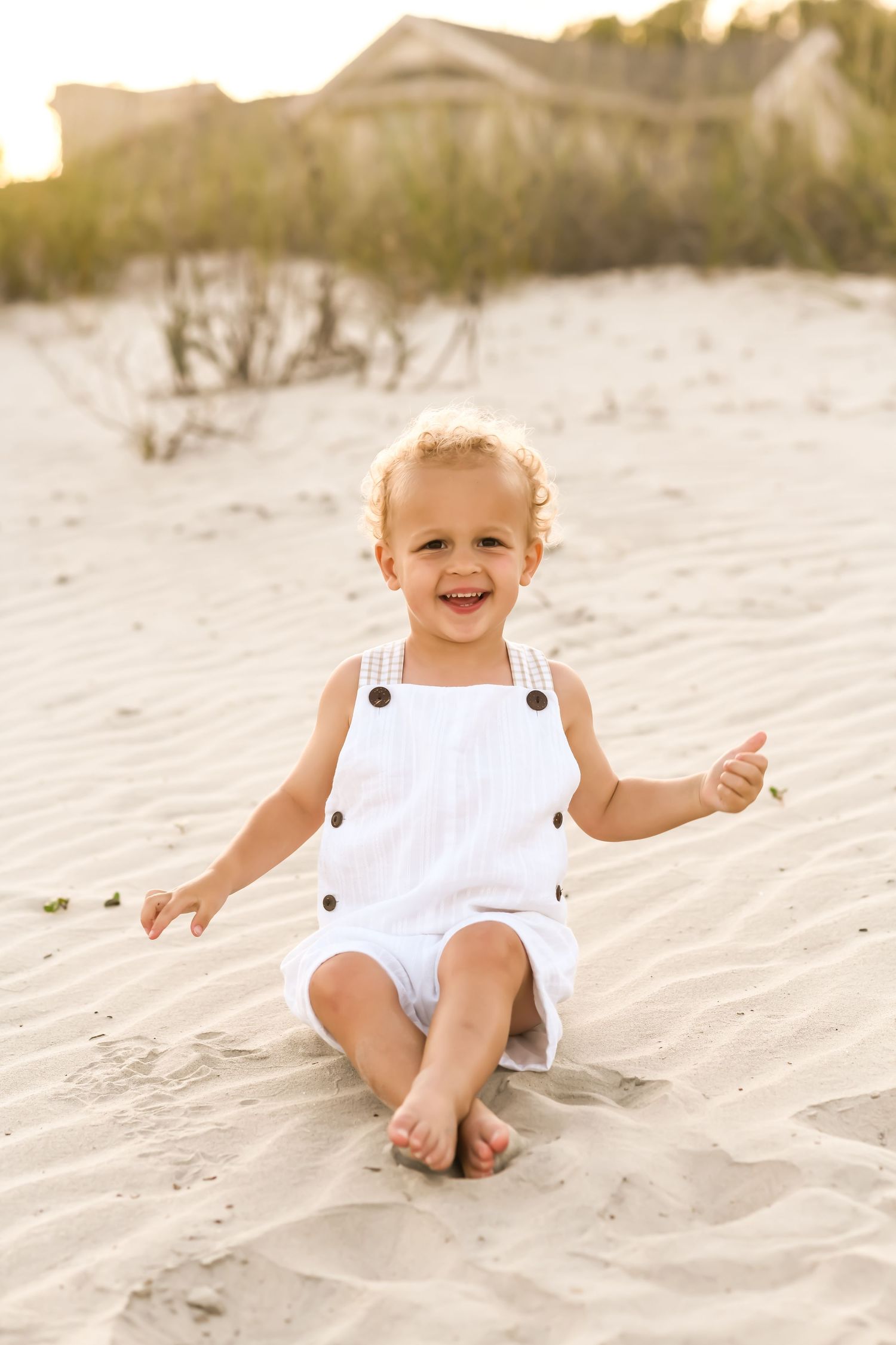 Young child wearing white overalls sitting and smiling on sandy beach at sunset on Hilton Head Island.