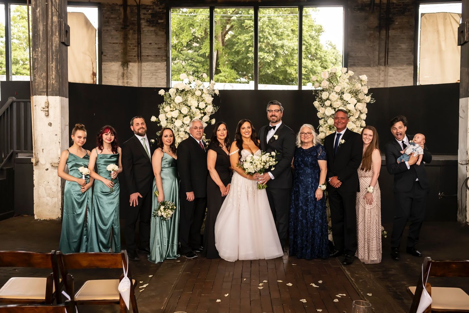 Wedding party poses together under floral decorations in industrial-style venue with wooden floors.