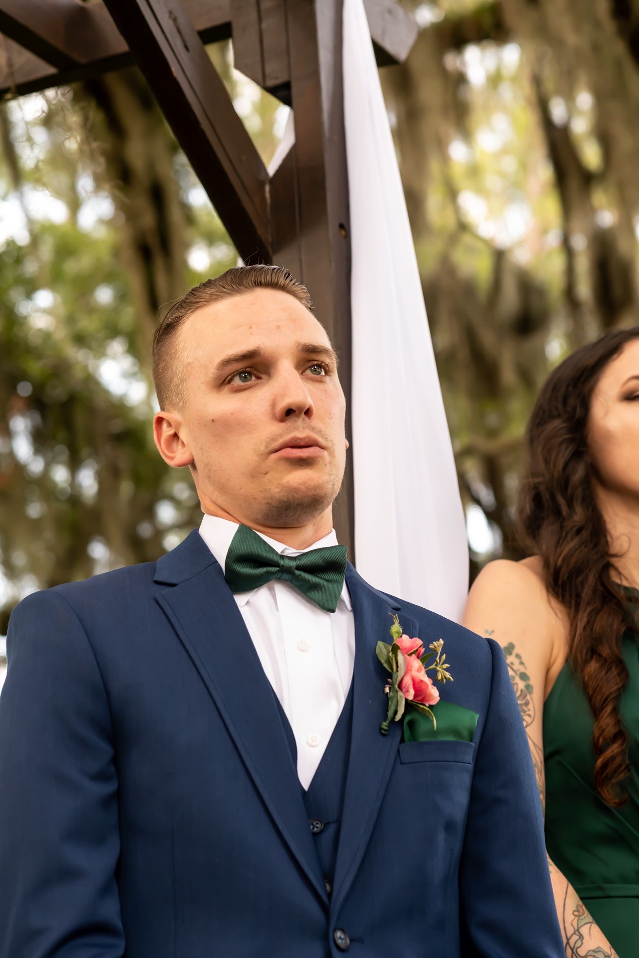 A groom in a navy suit with emerald green bow tie and floral boutonniere looks upward during an outdoor ceremony.