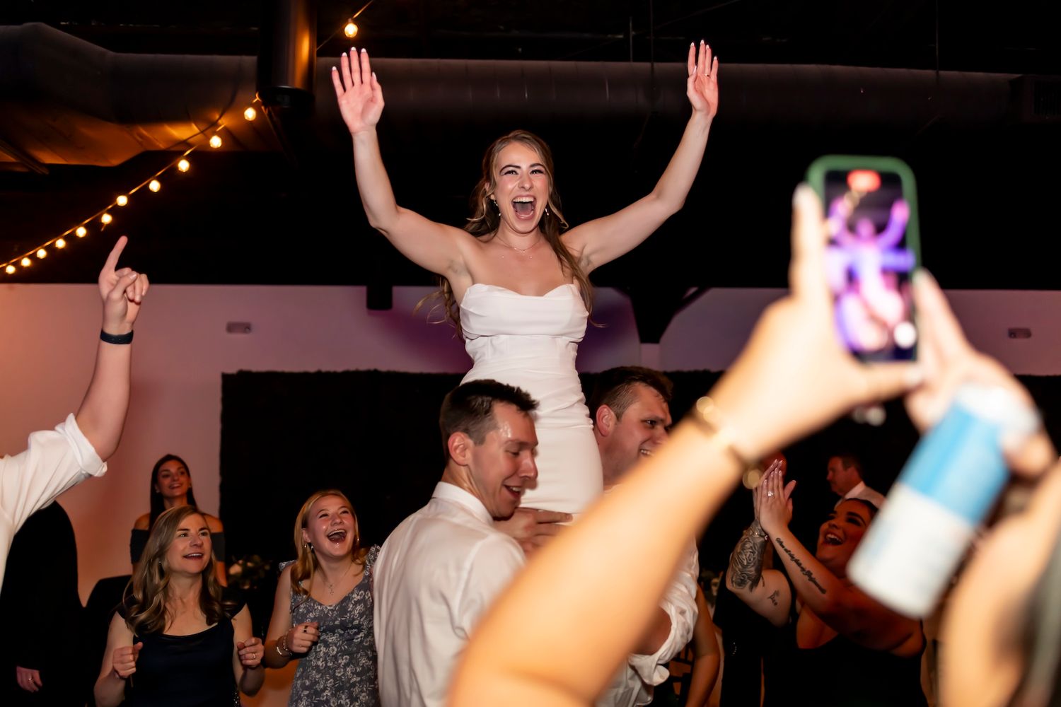 Wedding celebration with person being lifted high above the dance floor while other guests celebrate and take photos.