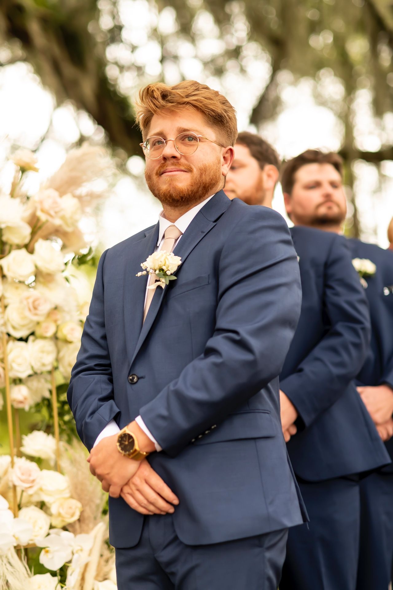 Groomsman in navy blue suit with floral boutonniere stands at outdoor wedding ceremony with white floral arrangements.