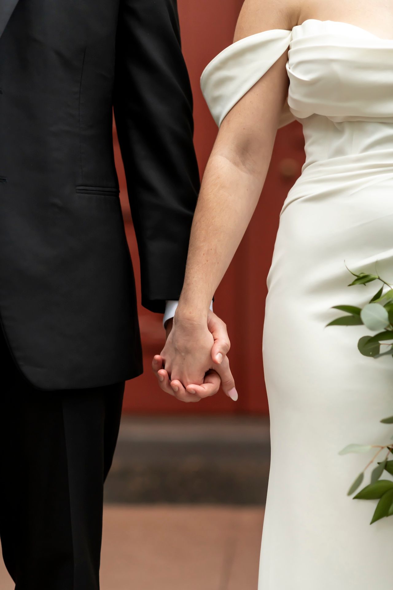 Close-up of a bride in white dress and groom in black suit holding hands against a reddish-orange wall background.