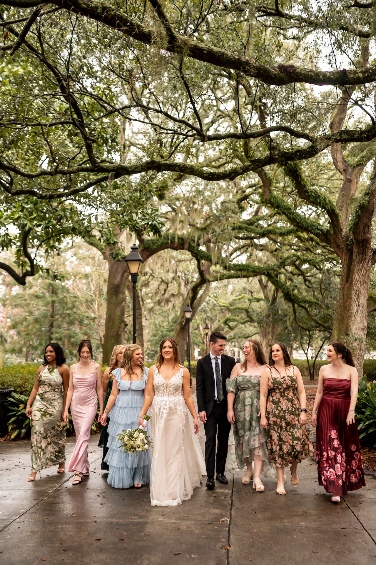 A wedding party walks together under moss-draped oak trees on a garden path wearing mix-and-match formal dresses.