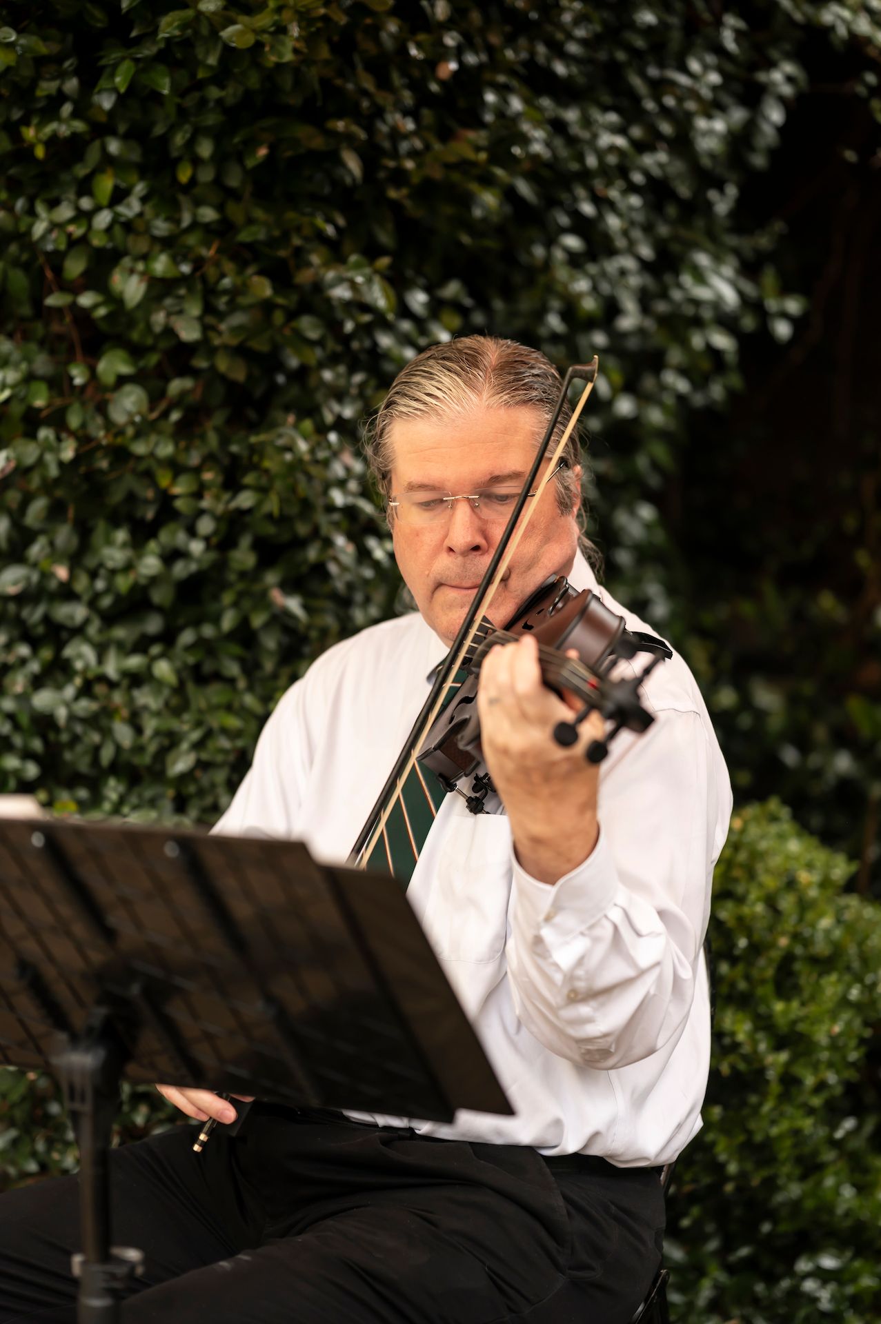A musician in white plays violin while reading sheet music at an outdoor performance.