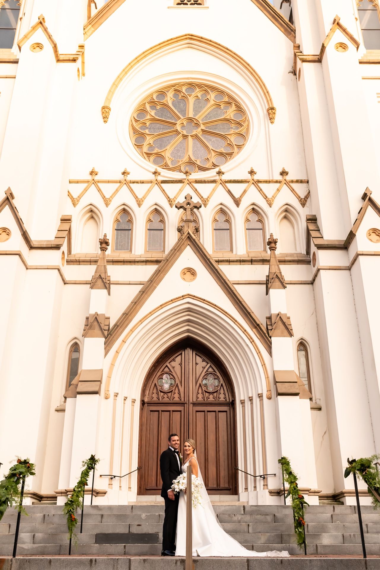 Elegant white Gothic church facade with arched doorways, rose window and stone steps adorned with greenery.