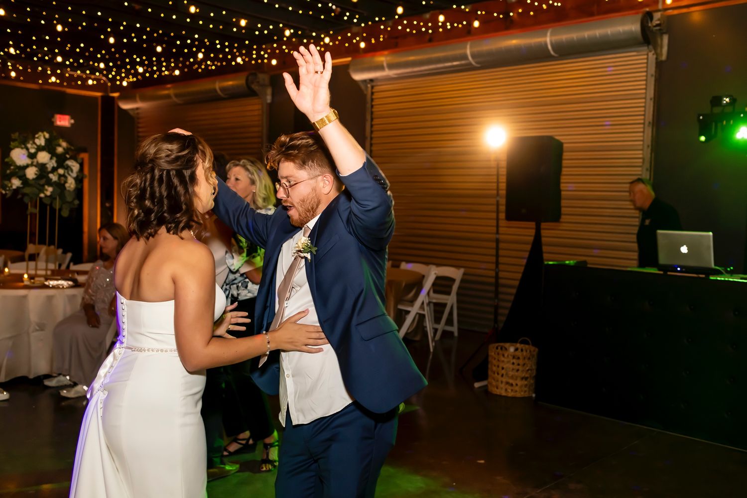 A group of wedding guests dancing at a festively decorated indoor reception venue with string lights overhead.