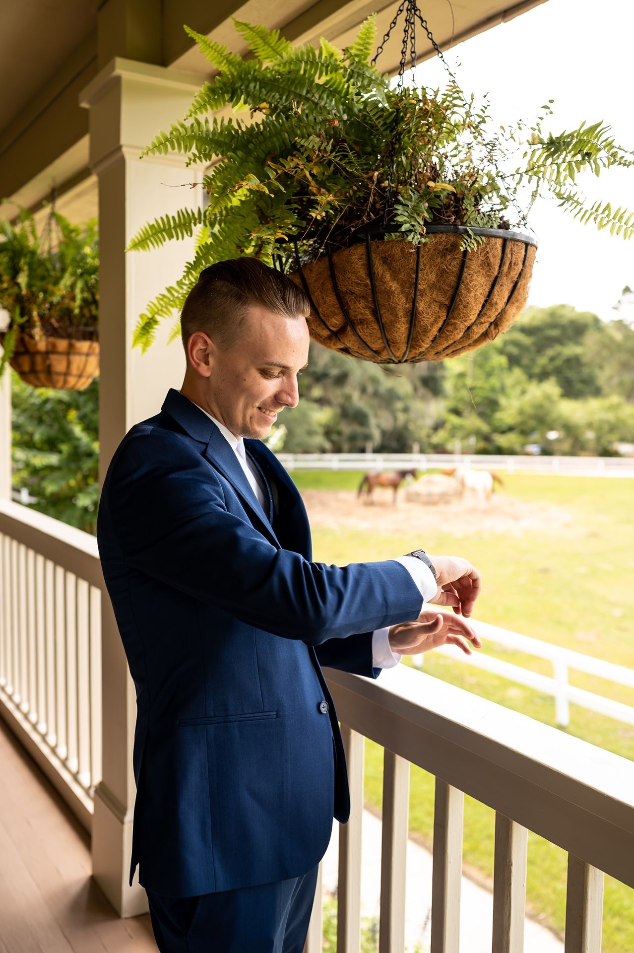 Person in navy suit adjusts cuff links while standing on a porch with hanging ferns and field views in background.