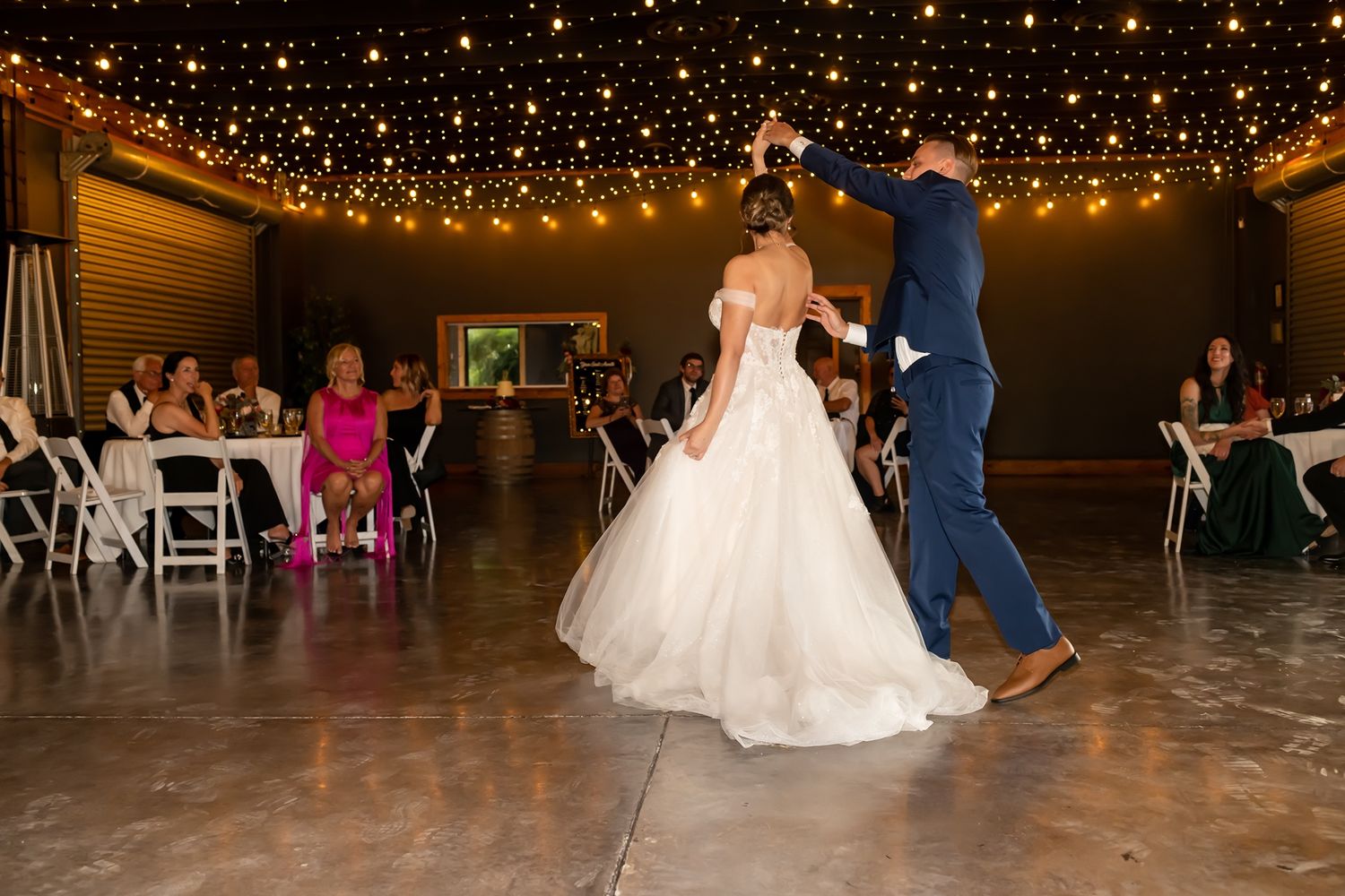 A romantic first dance at a wedding reception under twinkling string lights in a rustic venue space.