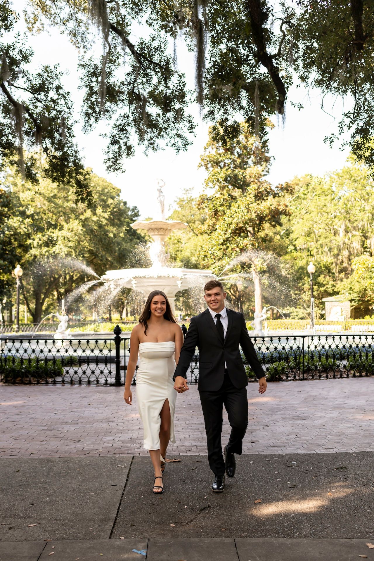 A couple walks together along a path in a scenic park with a beautiful ornate fountain in the background.