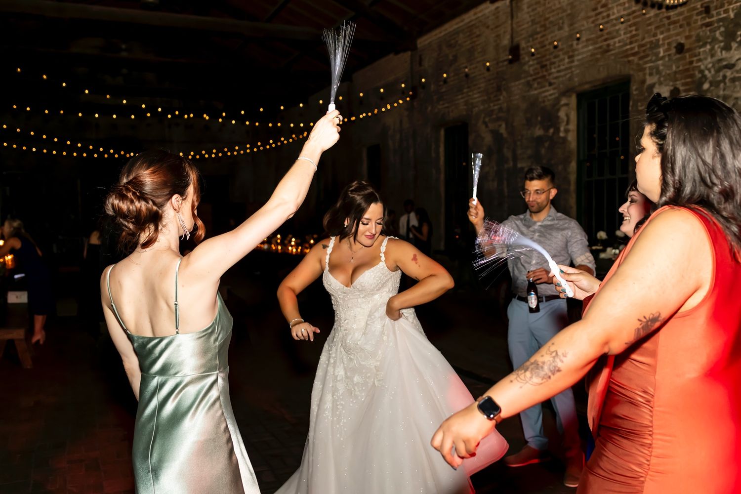 Joyful people dance and celebrate at a rustic wedding reception venue with string lights overhead.