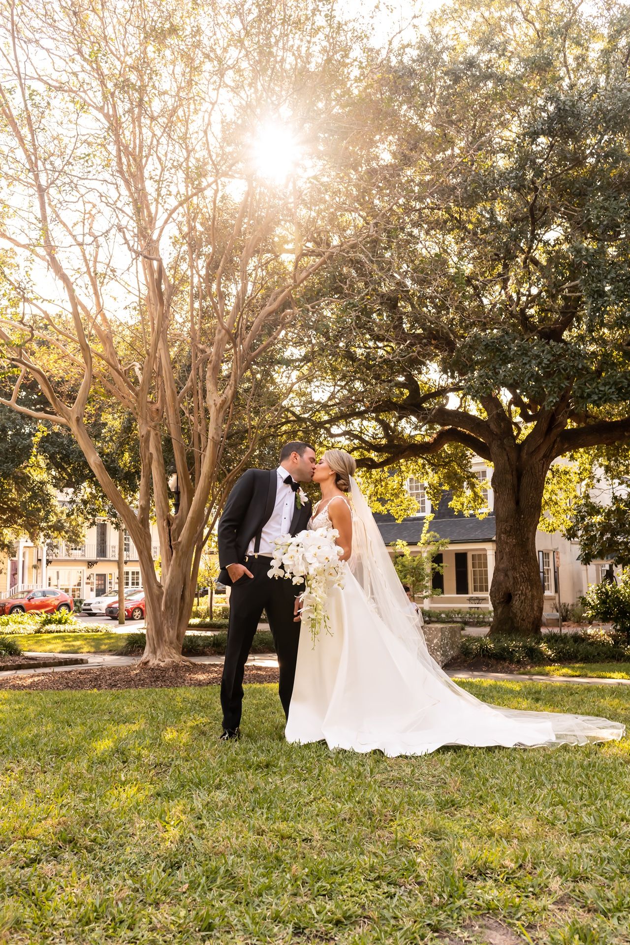 A newlywed couple shares a romantic moment in a sun-drenched garden with large oak trees at sunset.