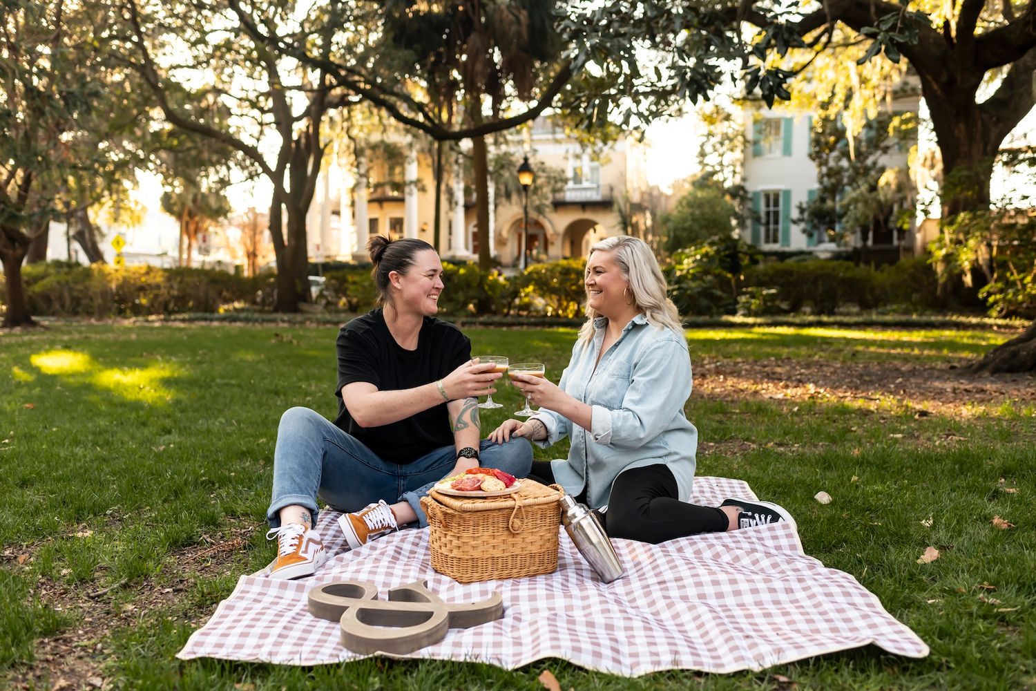 Two people enjoying a romantic picnic on a checkered blanket with a wicker basket in a sunlit park at golden hour.