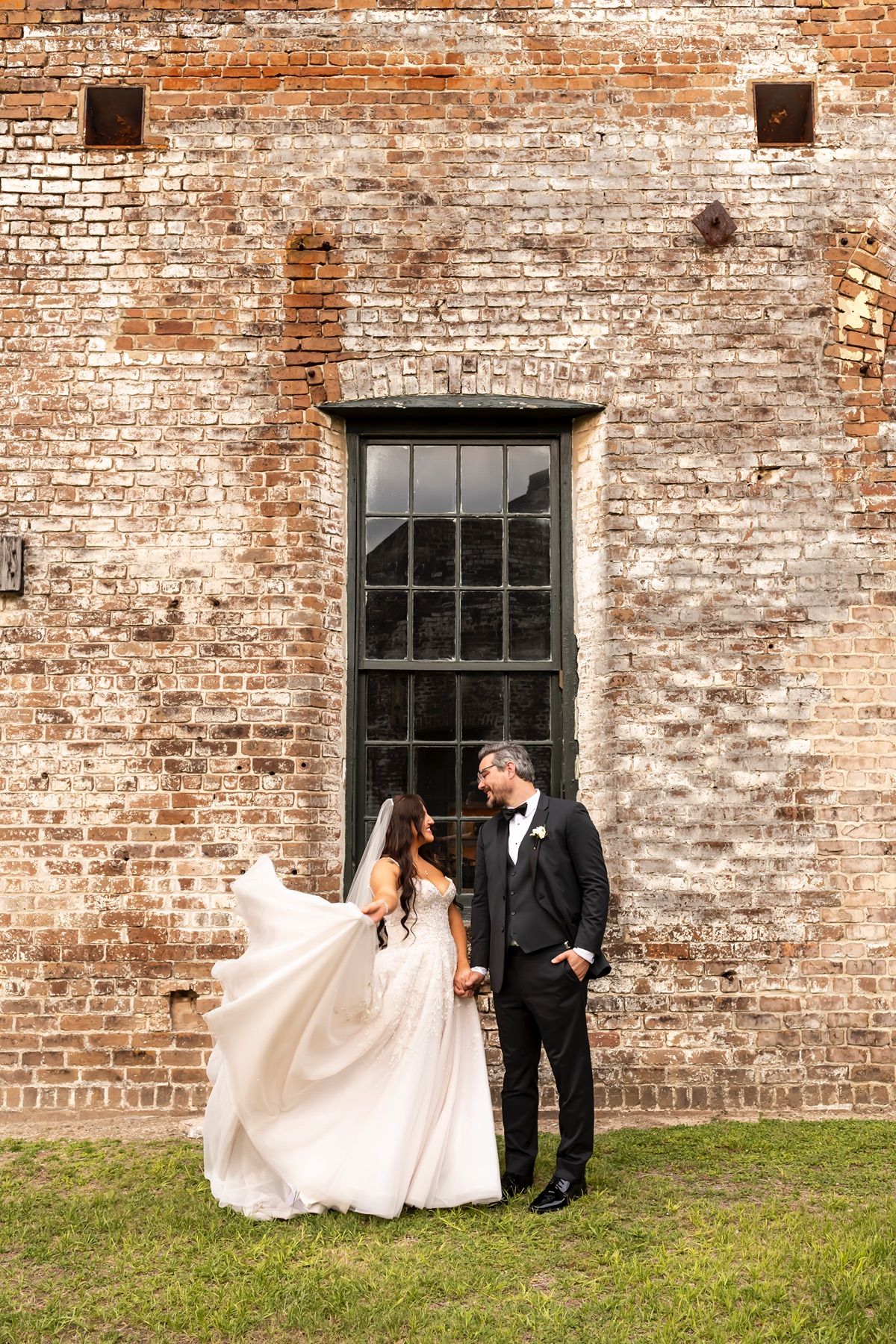 Two people in formal wedding attire walk hand in hand along a rustic brick wall during an outdoor photo session.
