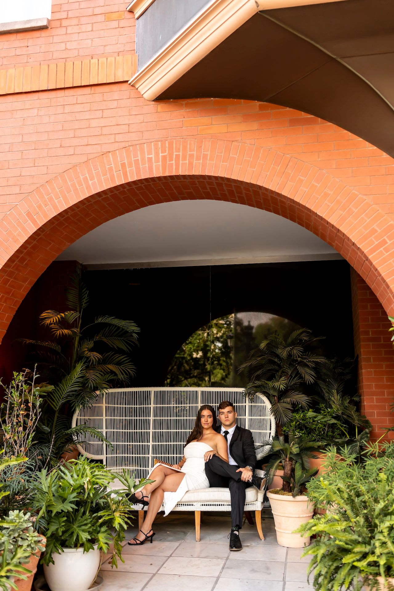 A covered outdoor seating area with brick arches, plants, and wooden chairs creating a cozy garden atmosphere.