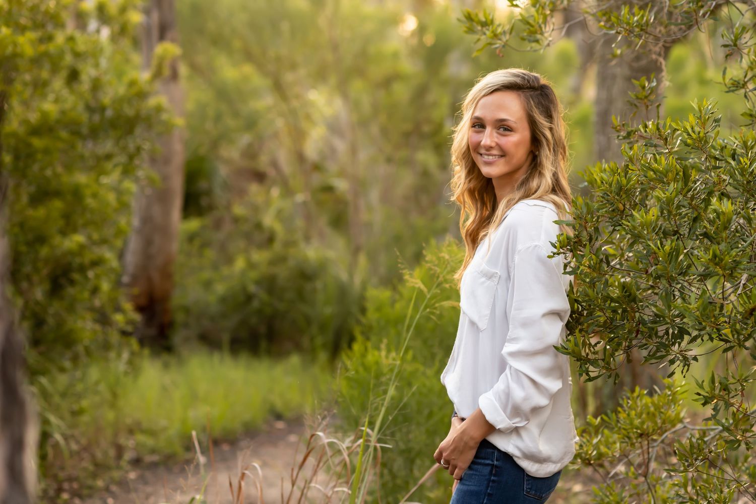 Person in a white sweater stands smiling in a lush natural outdoor setting with golden sunlight filtering through trees.