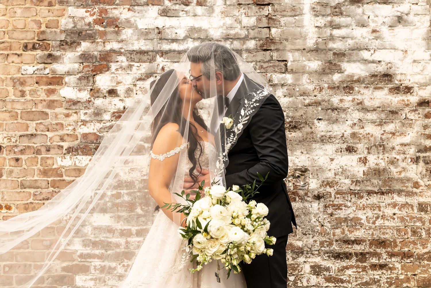 A romantic wedding couple embraces in front of a rustic brick wall while the bride holds a white floral bouquet.