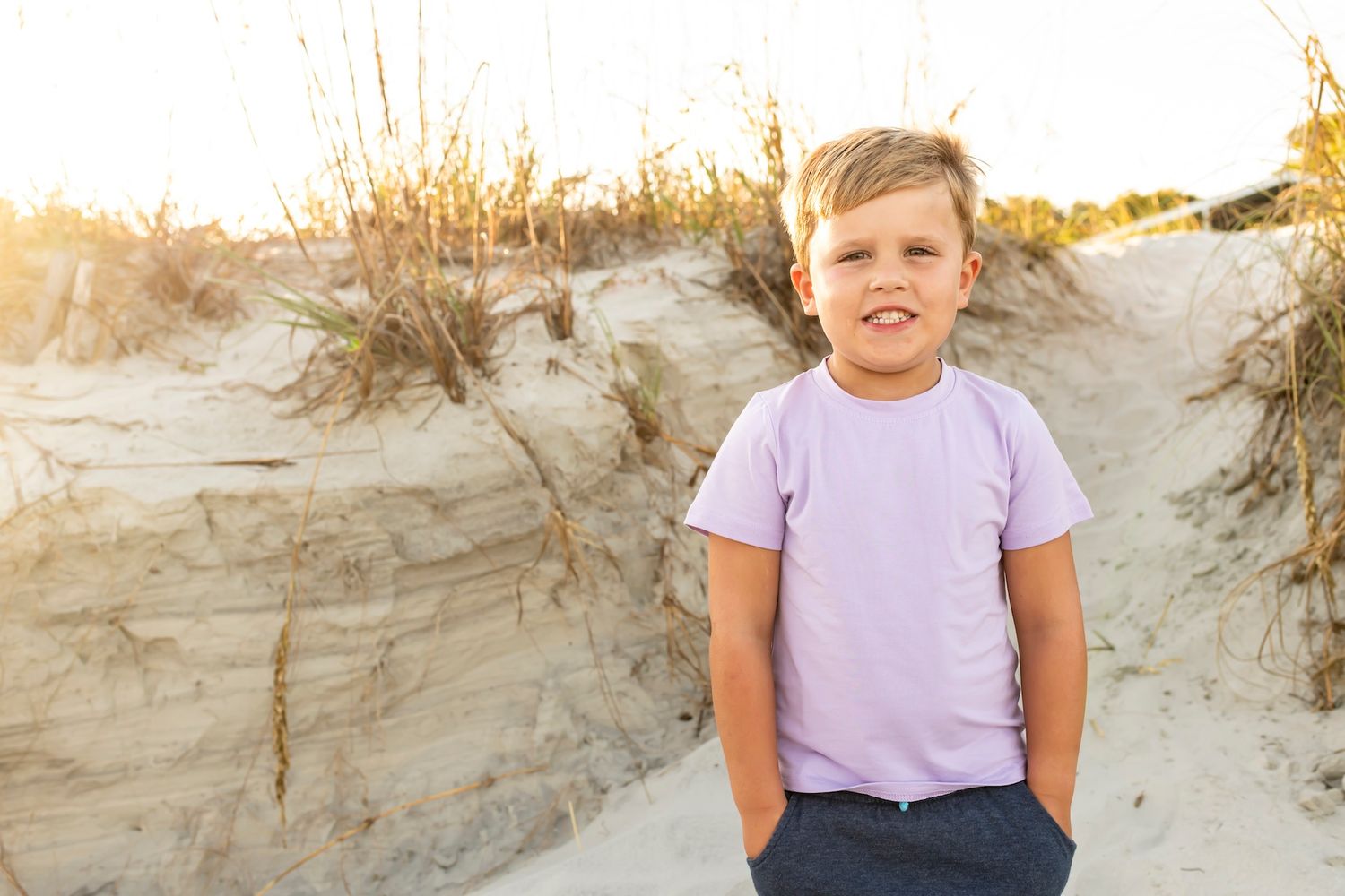 A child stands on a sandy beach dune during golden hour wearing a lavender t-shirt.