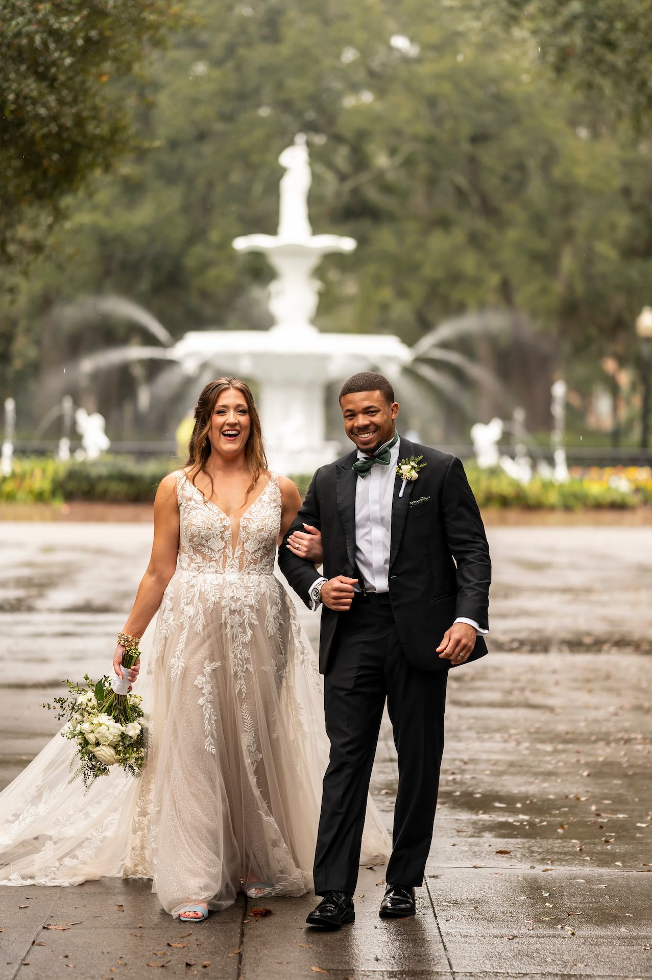 Newlyweds walking together on rainy day by ornate white fountain in garden setting.