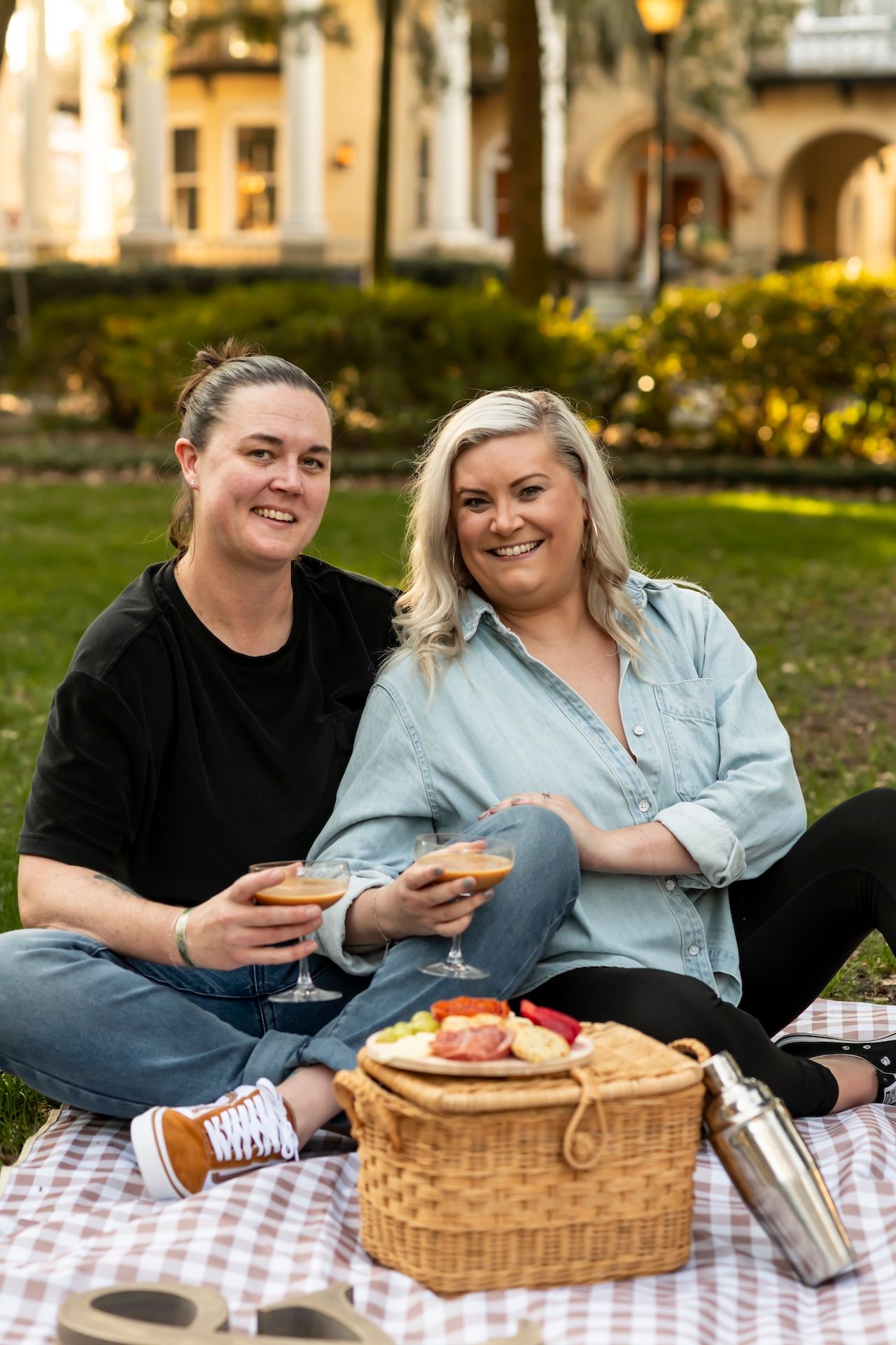 Two people enjoy a relaxing picnic with a wicker basket on a checkered blanket in a sunny garden setting.