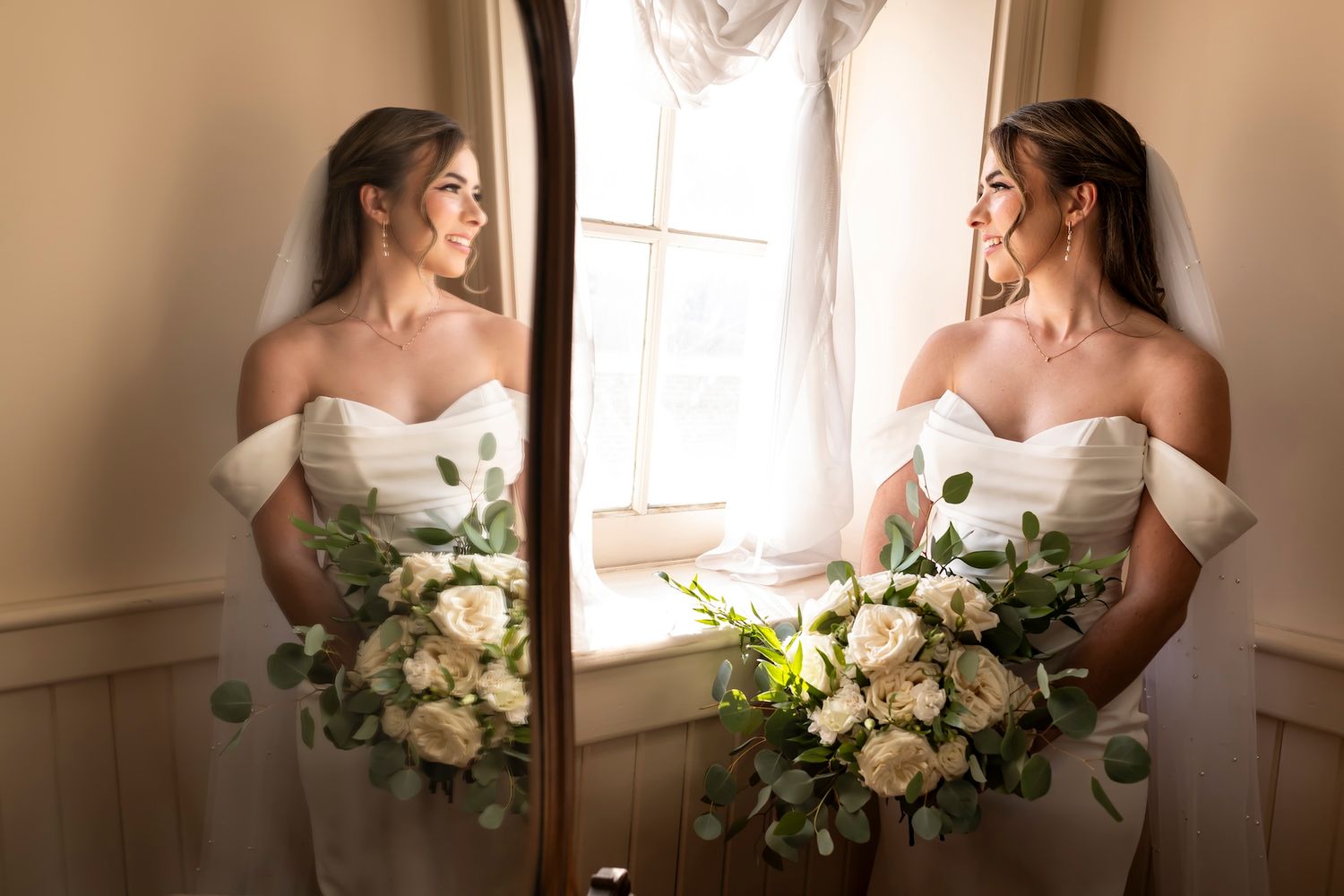 A elegant bride in a white off-shoulder gown holds a lush cream and green floral bouquet while standing by a window.