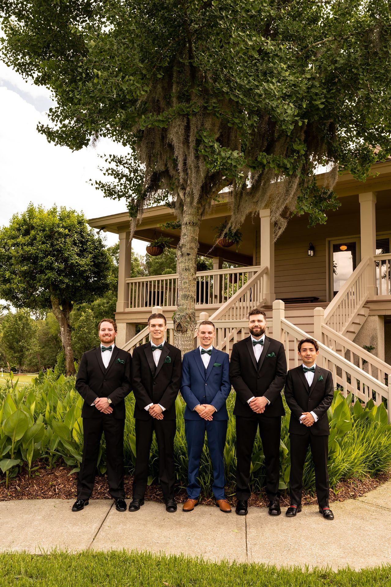 Groomsmen in formal attire stand in a row on a garden walkway in front of a house with white railings and tall trees.