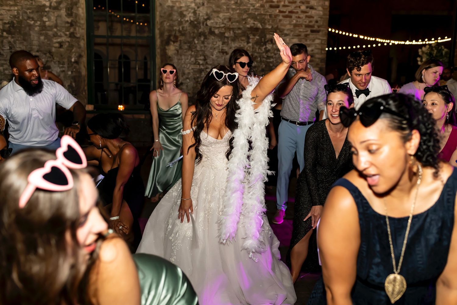 Wedding reception guests dancing and celebrating on the dance floor with string lights and exposed brick walls in the background.