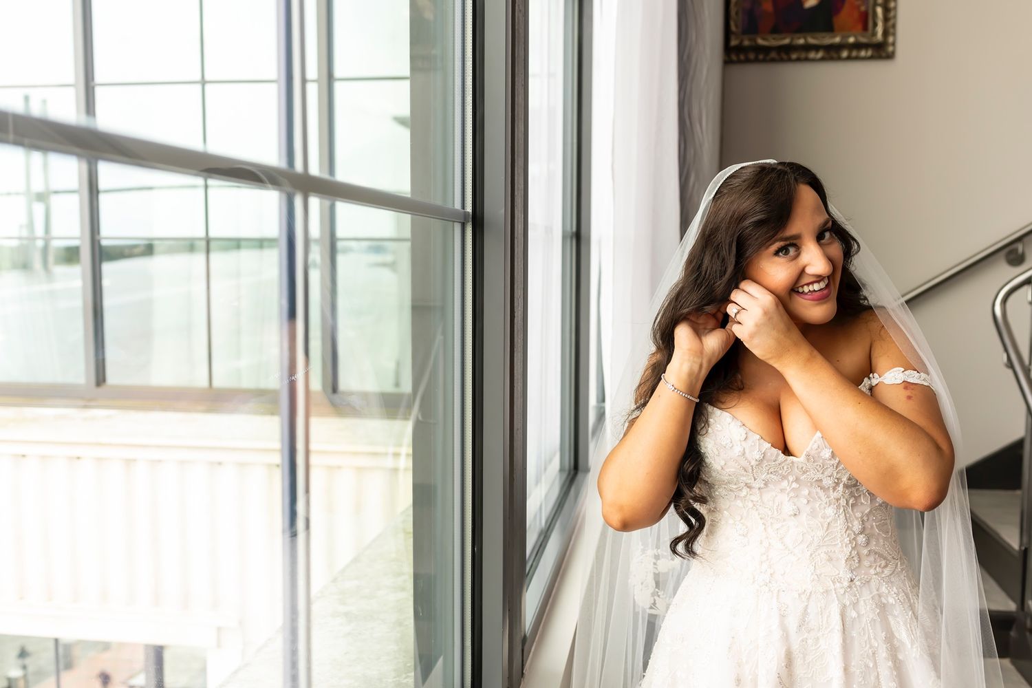 A bride in a white lace gown stands by a bright window while adjusting her long dark hair and veil.