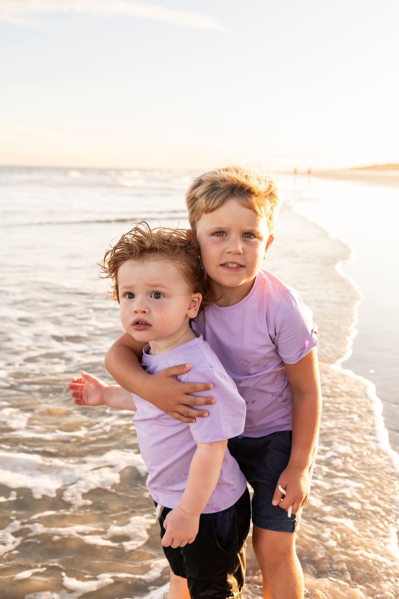 Two young children play and embrace on a sandy beach at sunset with golden light in the background.