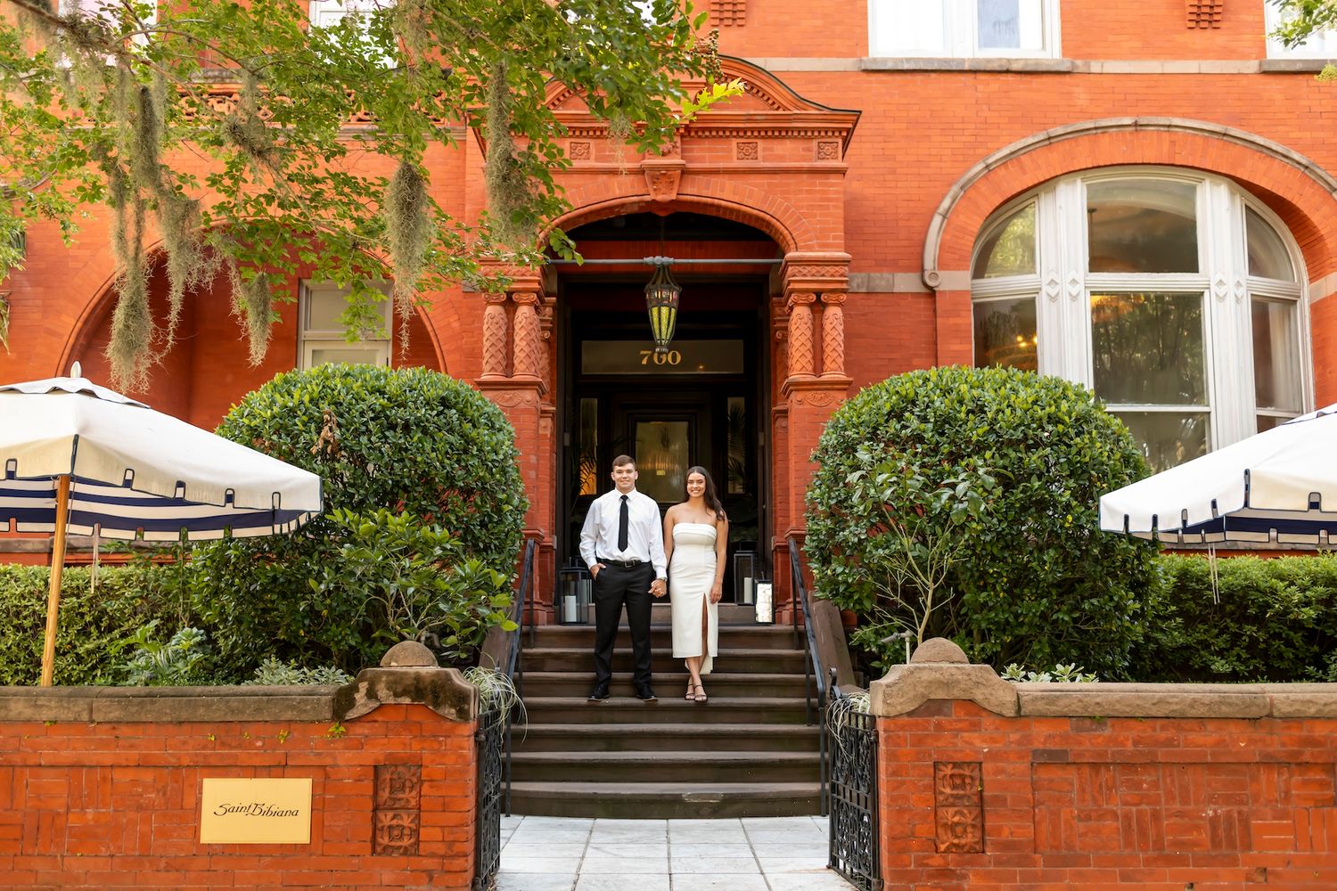Elegant red brick hotel entrance with stone steps, decorative bushes, and white patio umbrellas on either side.