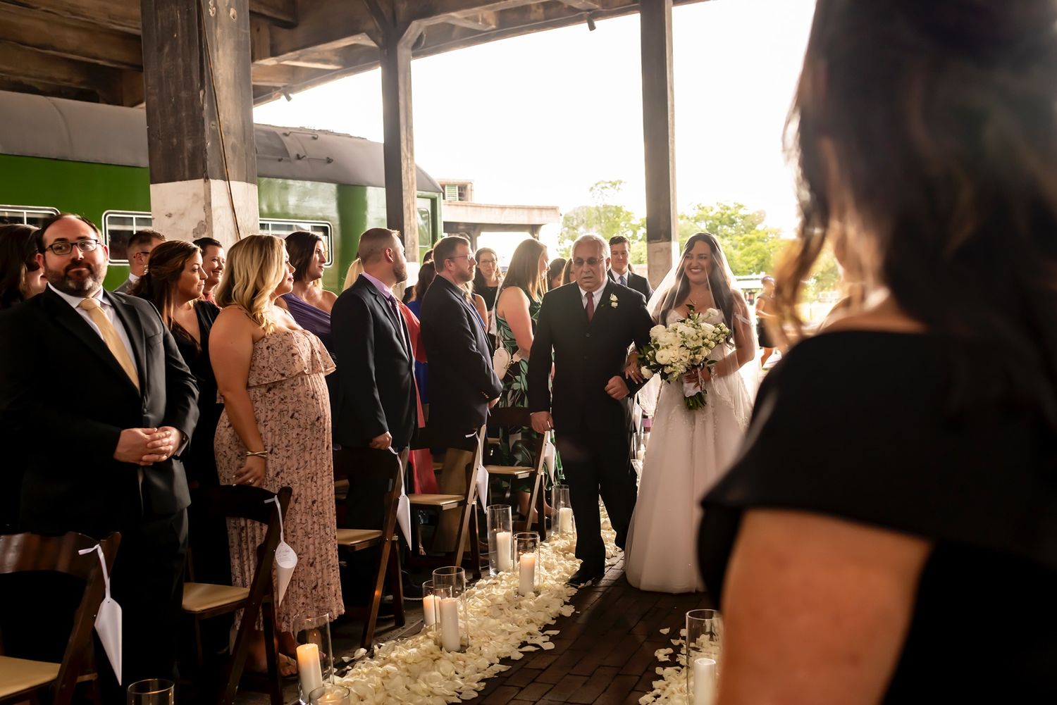 Wedding guests stand in rows for an outdoor barn ceremony as they watch a couple exchange vows at sunset.