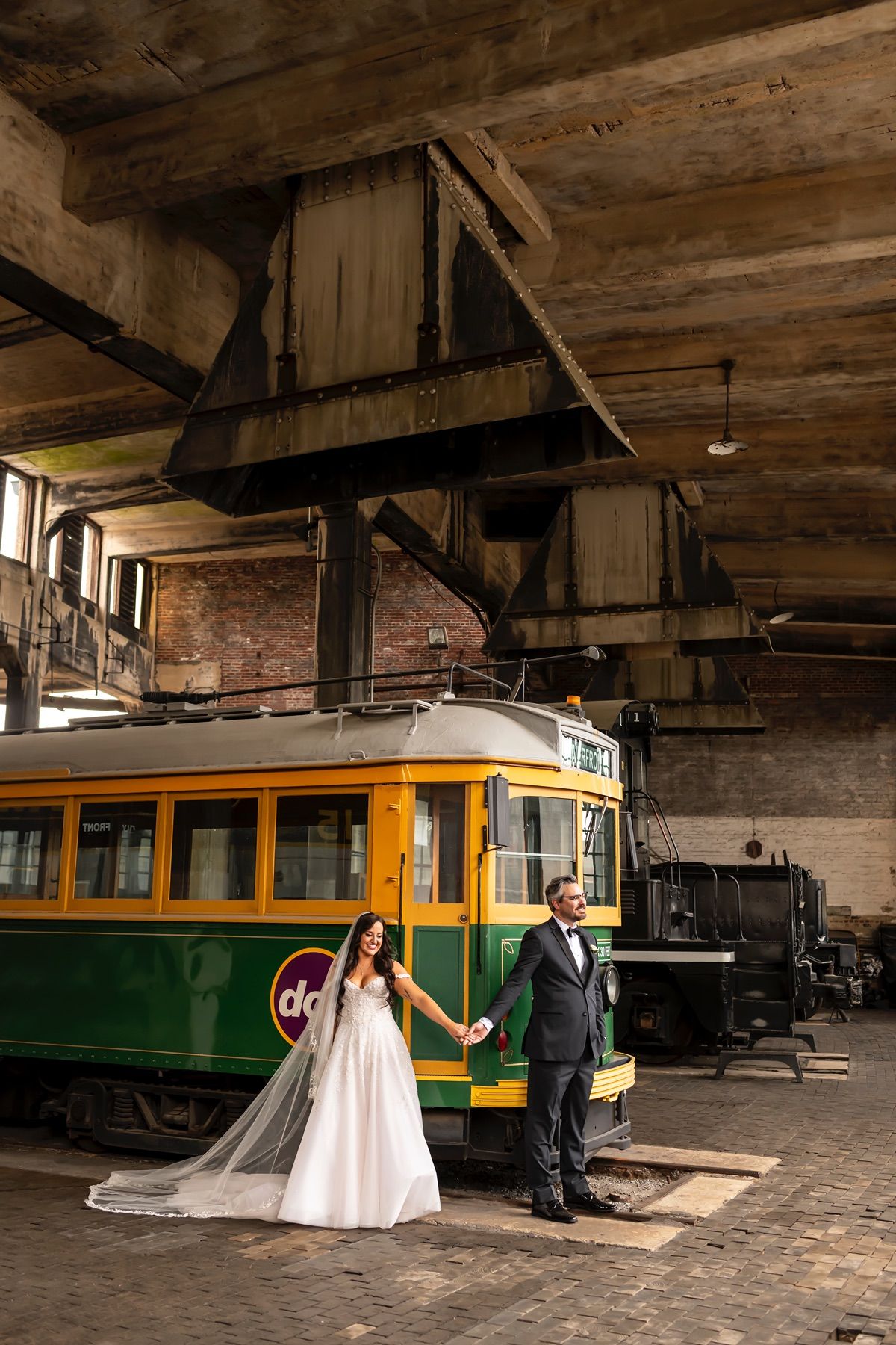 A couple in wedding attire poses romantically by a vintage green and yellow streetcar inside an industrial brick building.