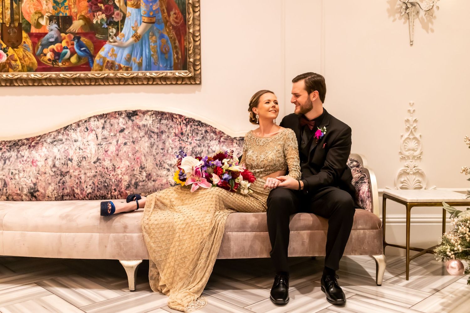Couple in formal attire relaxing on an elegant bench with soft warm lighting and floral decor in the background.