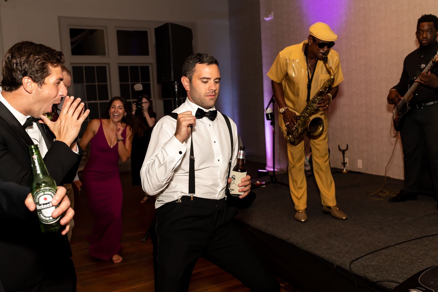 DJ performs energetically at a lively party while people dance in the dimly lit background under purple lighting.