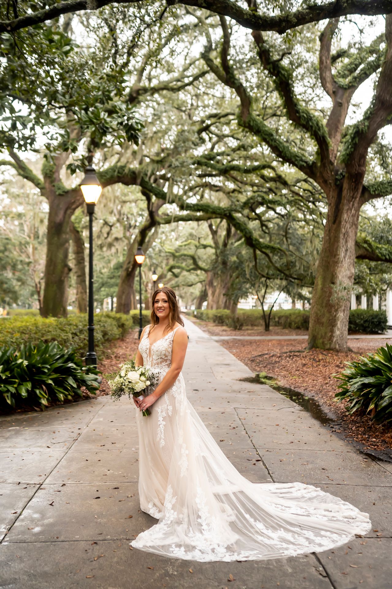A white lace wedding dress with long train photographed on a pathway lined with moss-draped oak trees in Savannah.